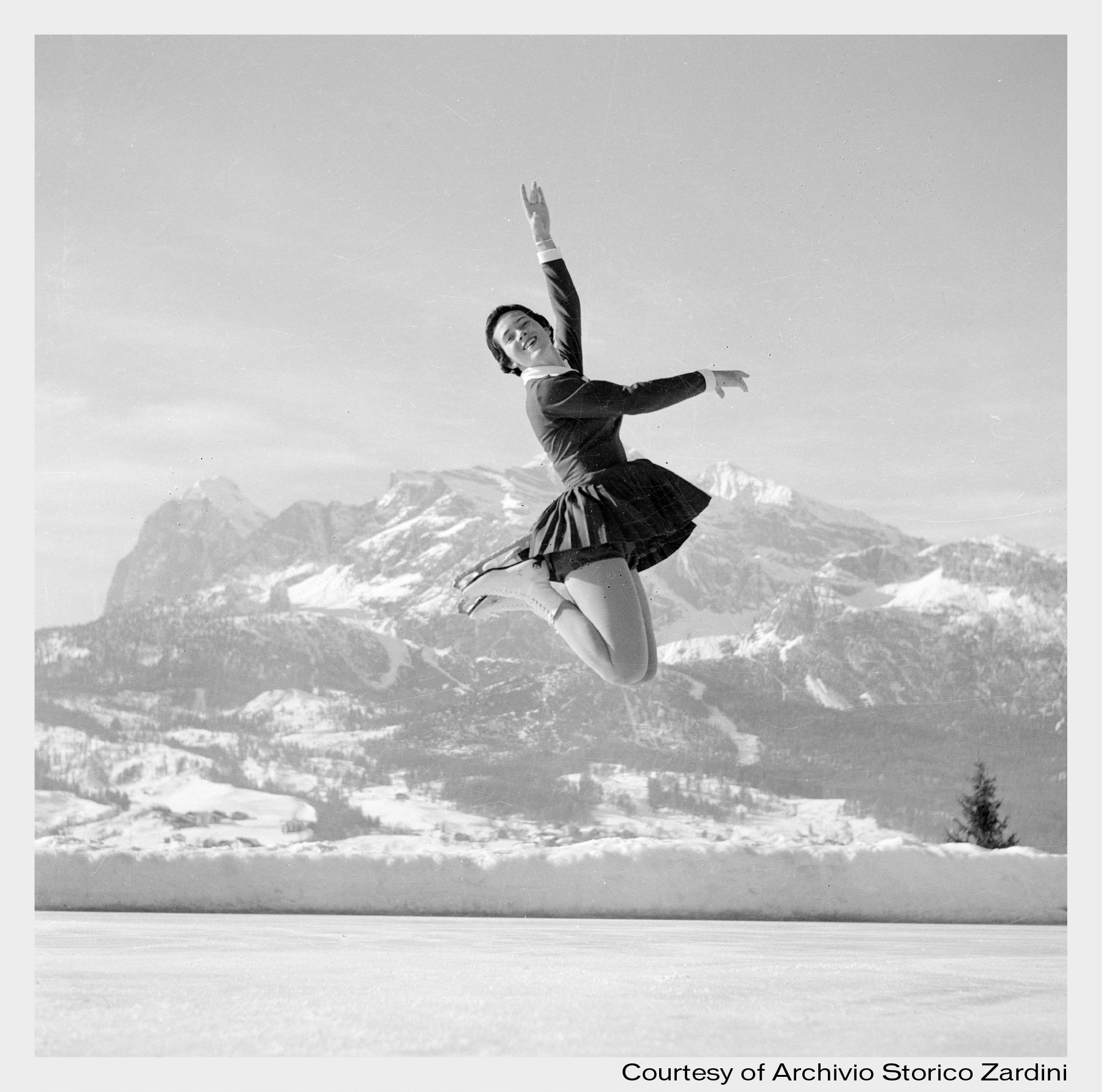 A female figure skater in a dress performs a graceful jump on an outdoor rink, snowy mountains and trees behind her under a clear sky.