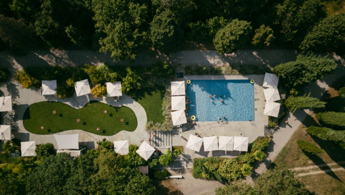 Aerial view of a pool with white umbrellas, lounge chairs, swimmers, and lush gardens with paths and more umbrellas nearby.