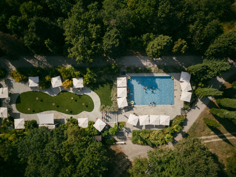 Aerial view of a pool with white umbrellas, lounge chairs, swimmers, and lush gardens with paths and more umbrellas nearby.