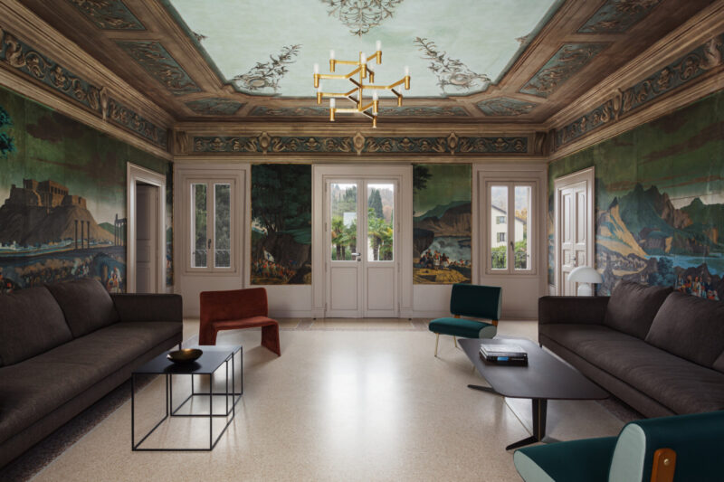 Elegant living room with mural walls, ornate ceiling, two dark sofas, modern tables, accent chairs, big windows, gold chandelier.