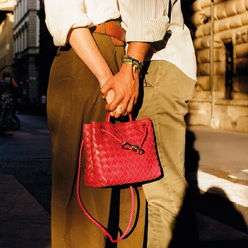 Two people stand close on a city street, holding hands behind backs. One carries a pink handbag. Sunlight casts shadows around them.