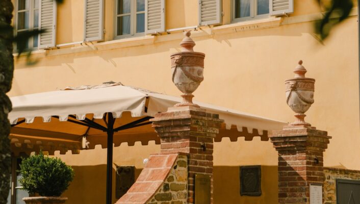 A sunlit courtyard with a stone wall, brick pillars crowned with urns, a potted plant, and a yellow-walled building featuring gray shutters. An awning provides shade for an outdoor seating area.