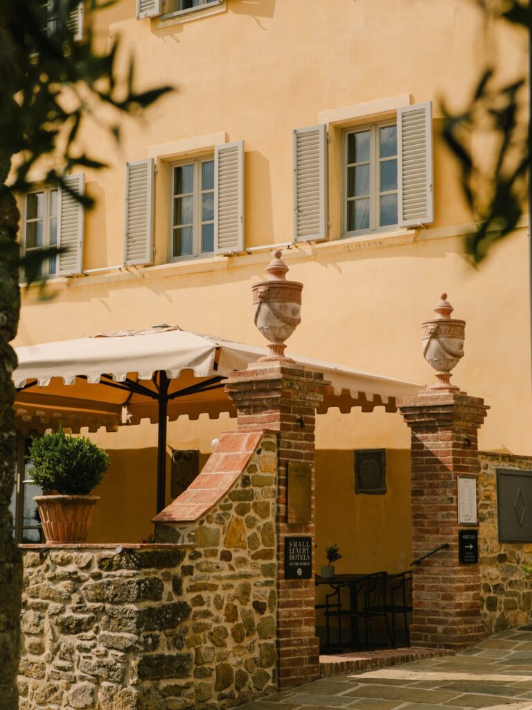 A sunlit courtyard with a stone wall, brick pillars crowned with urns, a potted plant, and a yellow-walled building featuring gray shutters. An awning provides shade for an outdoor seating area.