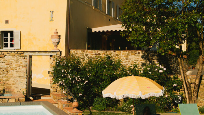 A person is relaxing beneath a yellow umbrella on a green lawn chair positioned near a swimming pool. They are next to a stone building featuring yellow walls and green shutters, all set amidst lush trees and blooming flowers.