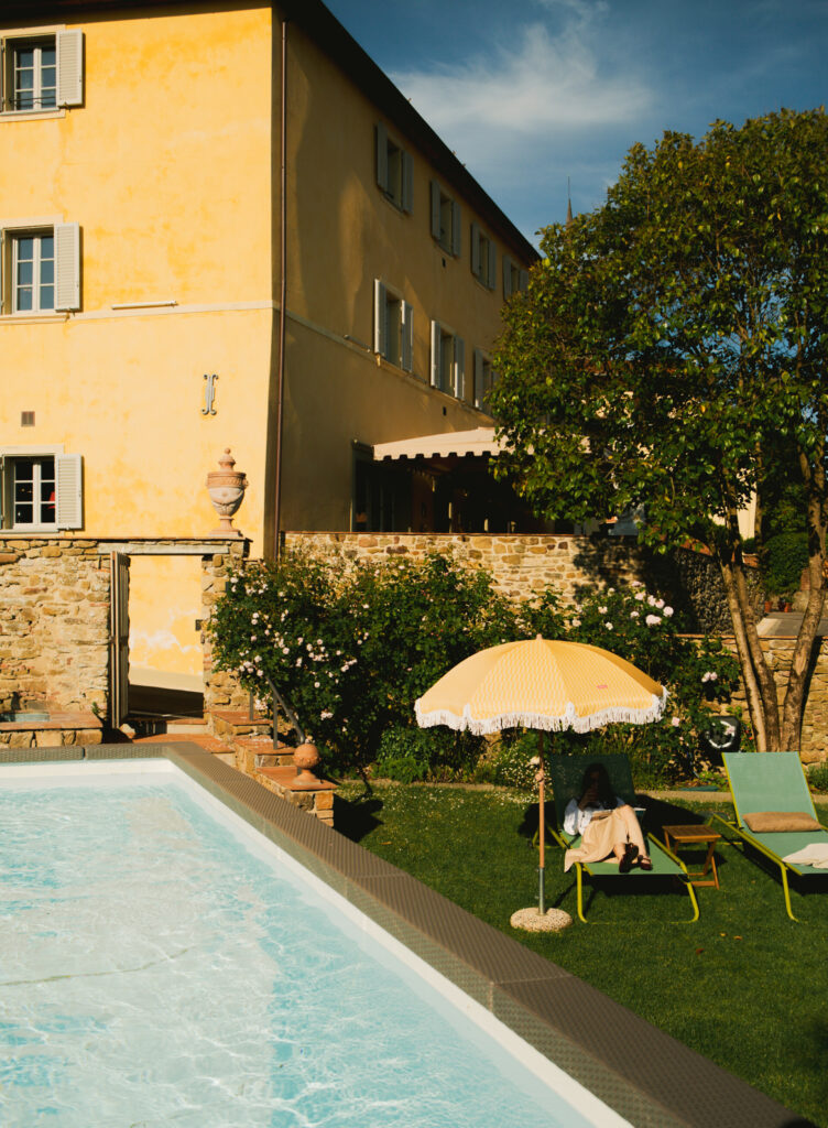 Una persona è seduta sotto un ombrello giallo accanto a una piscina, vicino a un edificio giallo e a un giardino rigoglioso, sotto un sole splendente.