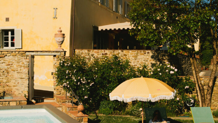 A person is relaxing beneath a yellow umbrella on a green lawn chair positioned near a swimming pool. They are next to a stone building featuring yellow walls and green shutters, all set amidst lush trees and blooming flowers.