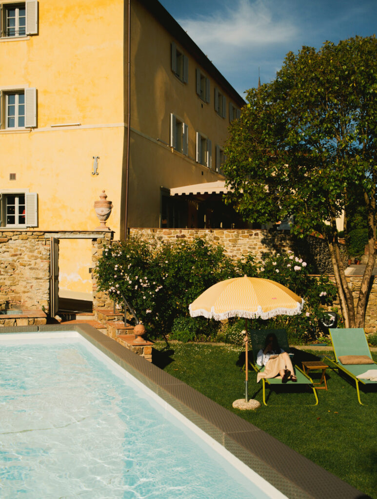 A person is relaxing beneath a yellow umbrella on a green lawn chair positioned near a swimming pool. They are next to a stone building featuring yellow walls and green shutters, all set amidst lush trees and blooming flowers.