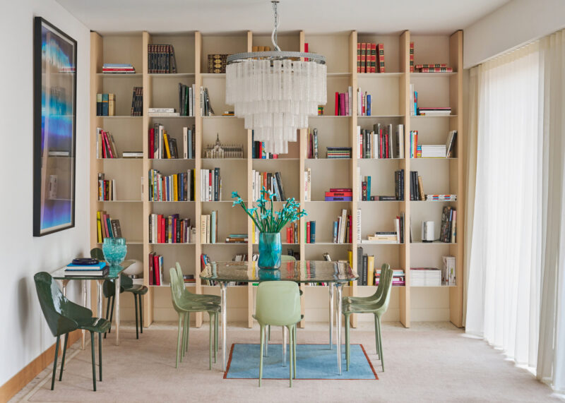 A modern dining room with a glass table surrounded by green chairs, a blue vase with fresh flowers as the centerpiece, an elegant chandelier overhead, tall bookshelves packed with books spanning the wall, and expansive windows draped in sheer curtains that bathe the space in soft natural light.