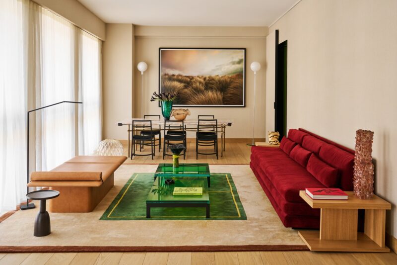 Modern living room with a red sofa, tan bench, glass coffee table, dining table featuring black chairs, large landscape artwork on the wall, and expansive floor-to-ceiling windows draped in light curtains. The space is decorated in neutral tones with pops of green accents.